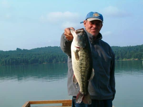 An angler holding a largemouth bass with a scenic view of hagg lake behind.