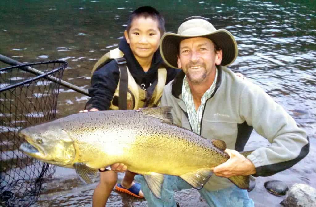 A man holding chunky spring chinook salmon caught fishing on the Rogue River, with a smiling boy behind the fish.