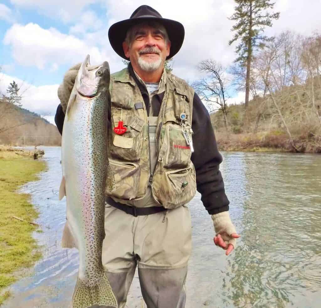 A man standing on the bank holding up a nice steelhead.
