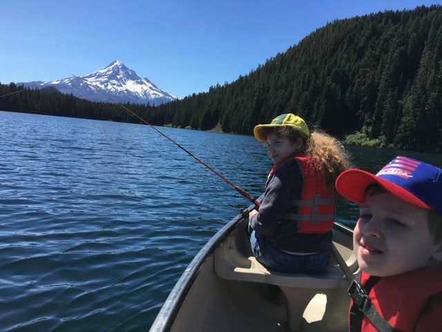 Two kid fishing on a boat with a scenic view of a lake and a mountain behind.