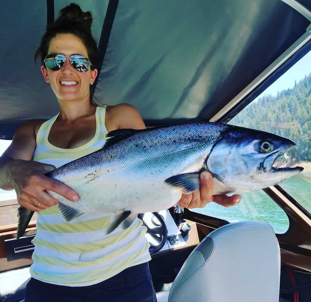 A smiling woman in a boat holds a giant landlocked chinook salmon caught fishing on Trinity Lake.