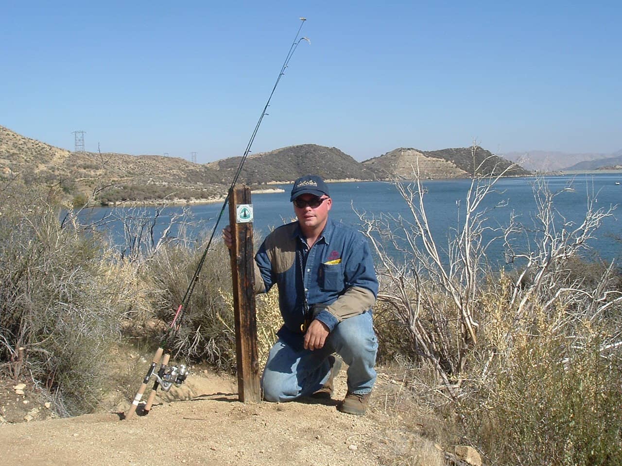 An angler with fishing pole kneels near a sign along the Pacific Crest Trail with Silverwood Lake in the background.