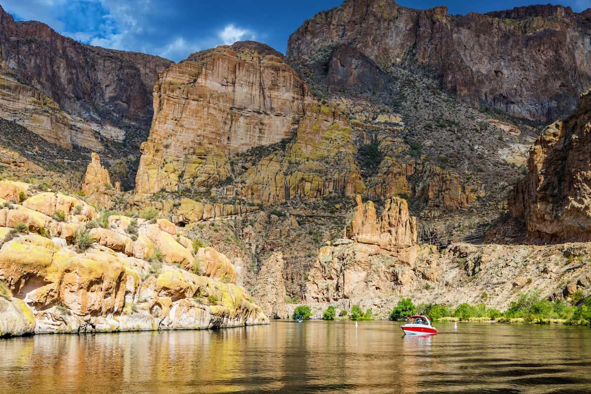 A red boat motors in front of yellow and brown cliffs at Canyon Lake, Arizona.