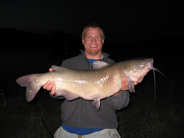 A man holds up a large catfish caught in the Missouri River in Montana, with a black night background.