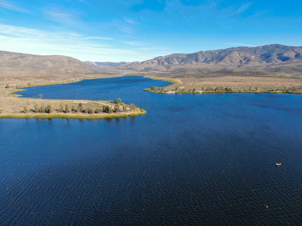 Aerial view of Lower Otay Lake (Reservoir) with blue sky and dry mountains in the background, and a small fishing boat on the blue water.