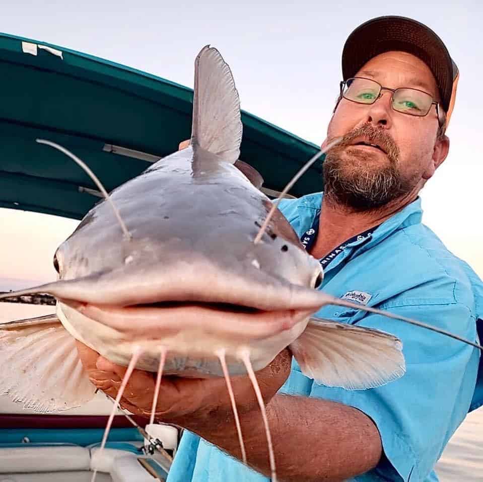 Man holds catfish facing directly into the camera with boat in background.