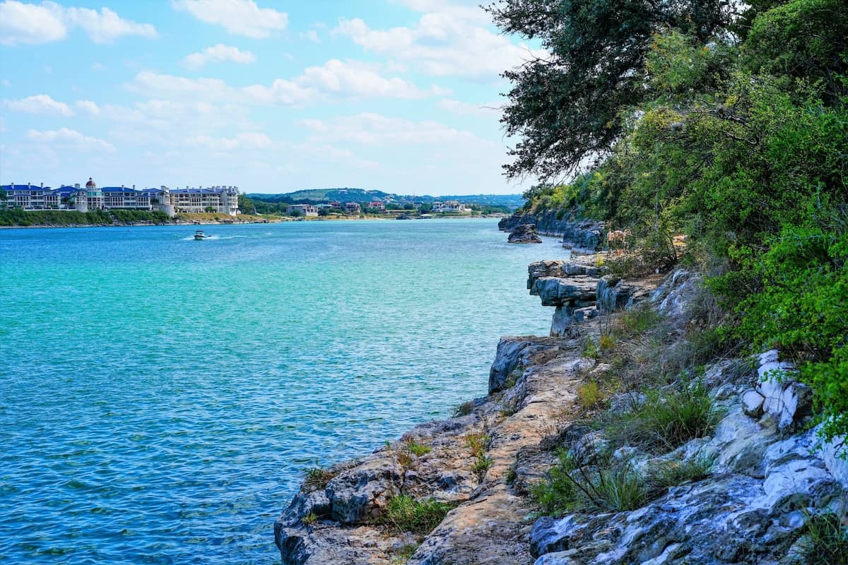 The rocky shoreline of Lake Travis in the foreground with a fishing boat speeding across the blue water in the distance.