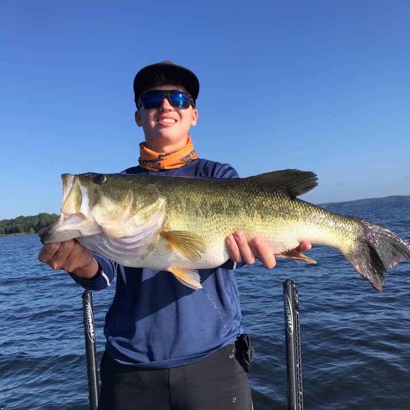 A young man in sunglasses holds a trophy bass he caught fishing in Toledo Bend Reservoir, with the lake water behind him.