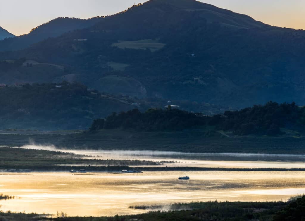 A fishing boat out on the surface of Lake Casitas as the sun sets with mountains in the background.