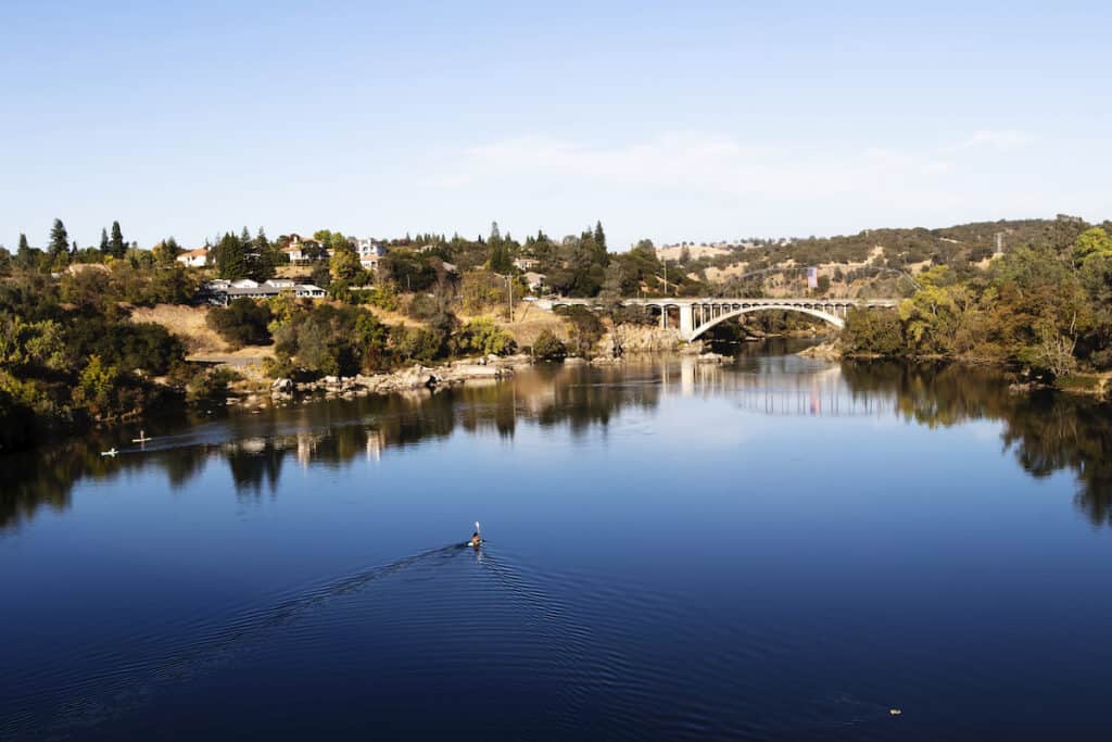 Lake Natoma's blue water with the Rainbow Bridge in the background and people paddling in the water.