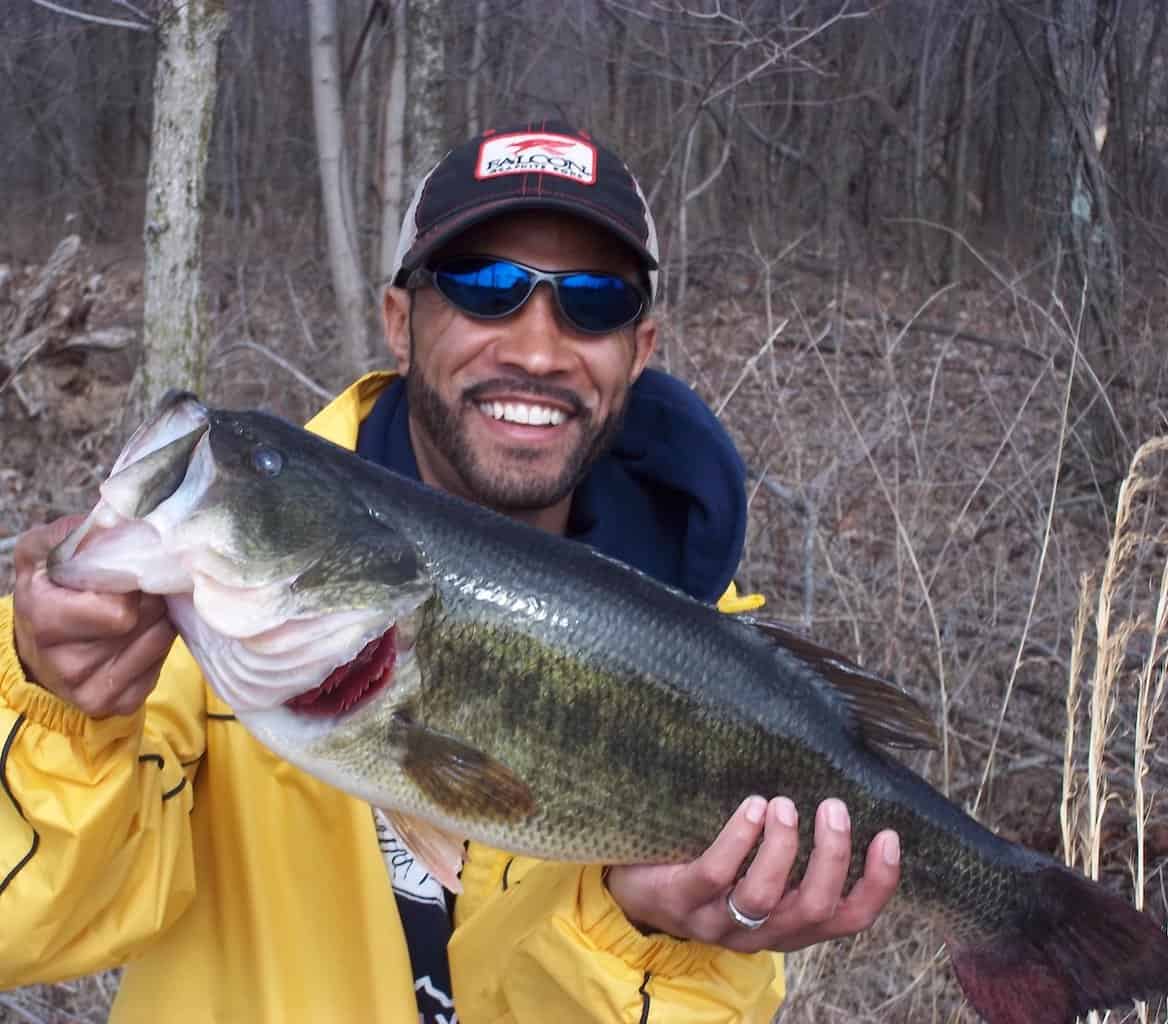 A man holds a big largemouth bass caught while fishing in Ohio, with woods in the background.