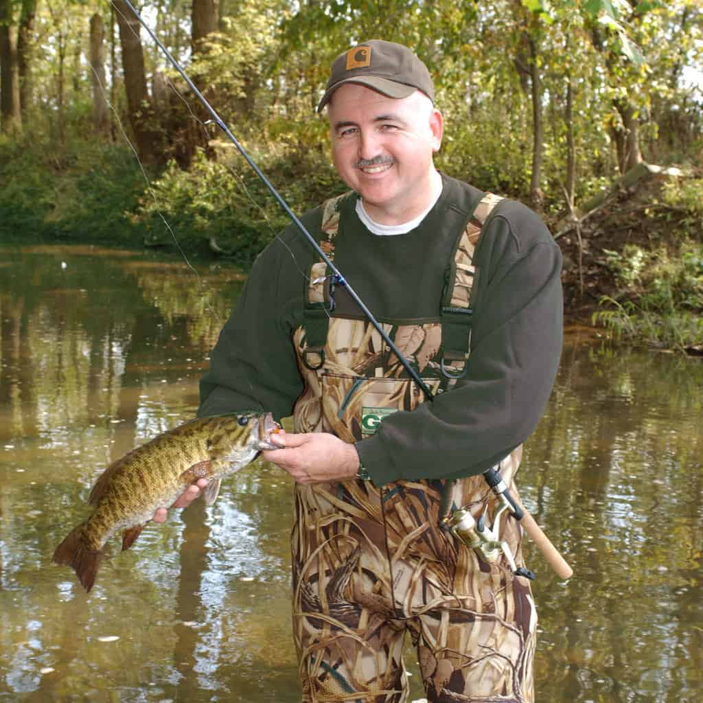 An angler in camouflage waders holds a smallmouth bass caught in an Ohio stream.