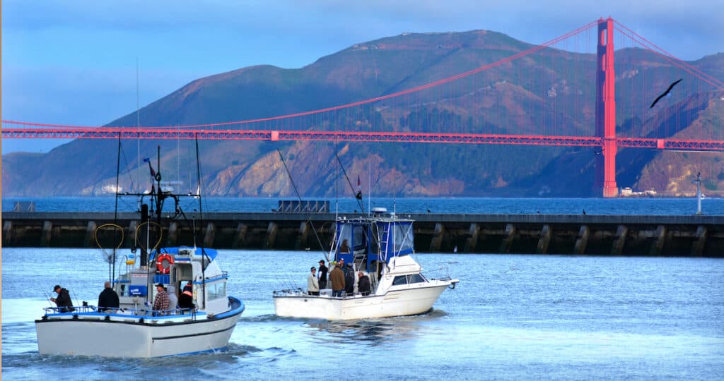Fishing boats sail out of Fisherman's wharf with the Golden Gate Bridge catching rays of sunrise in the distance.