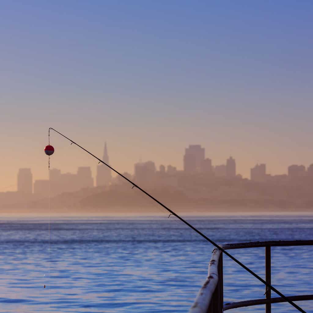 A fishing pole rests on a pier with the skyline of San Francisco in the background.