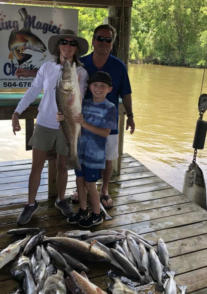 A man and woman with a boy hanging on to a large redfish along with a dock piled with more fish they caught in Louisiana.