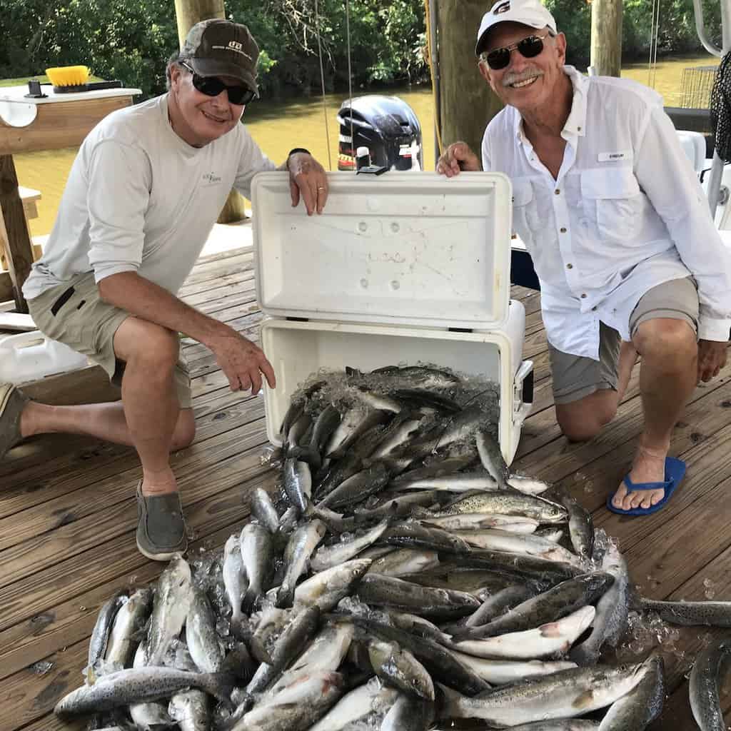 Two anglers kneeling next to a cooler spilling out dozens of speckled trout caught fishing in Louisiana.