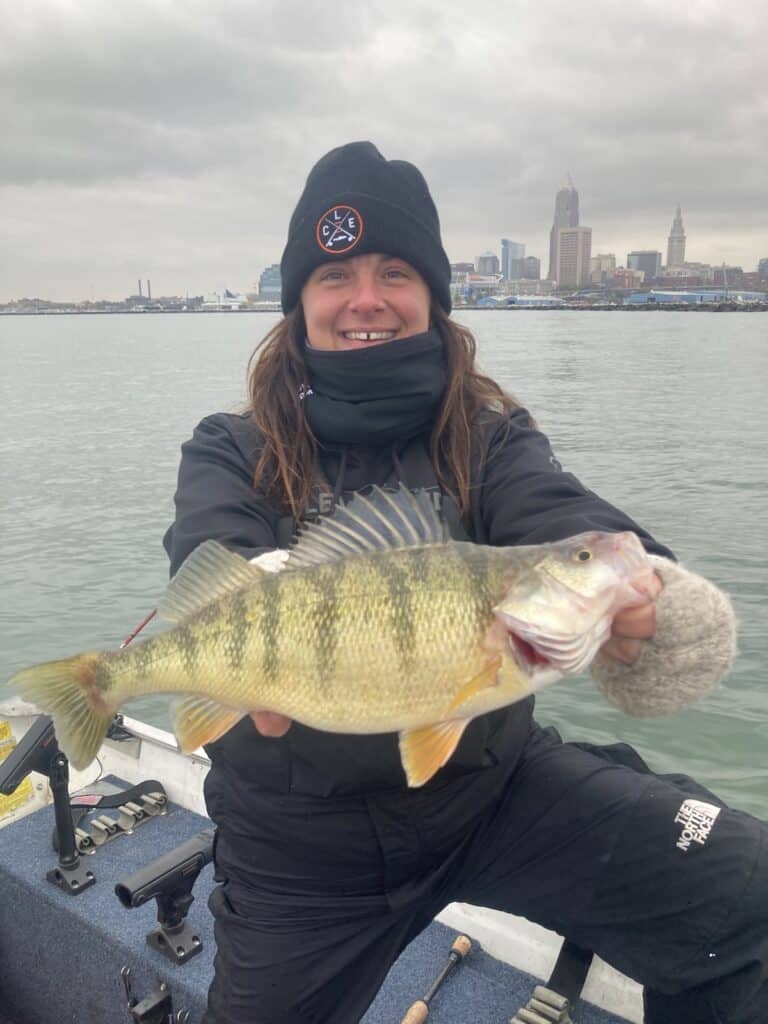 A woman in a fishing boat on Lake Erie holds a very large yellow perch, with Cleveland's skyline in the background.