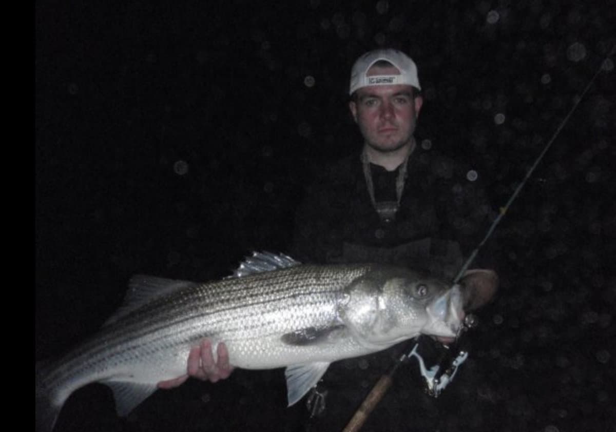 An angler in the pitch dark of night holds a larger striped bass (a.k.a. striper) caught fishing in Massachusetts.