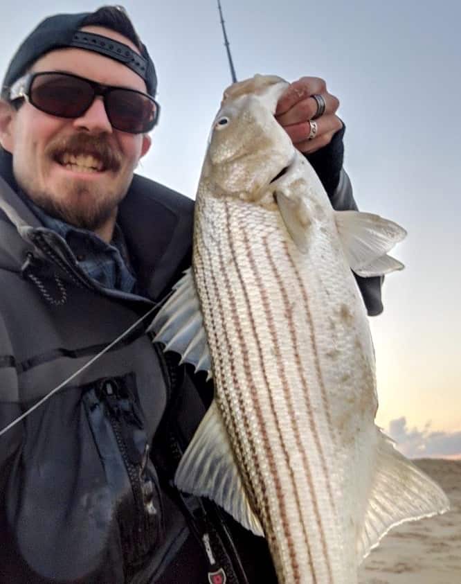 Angler holds up a striped bass in a selfie photo along the Atlantic coastline.