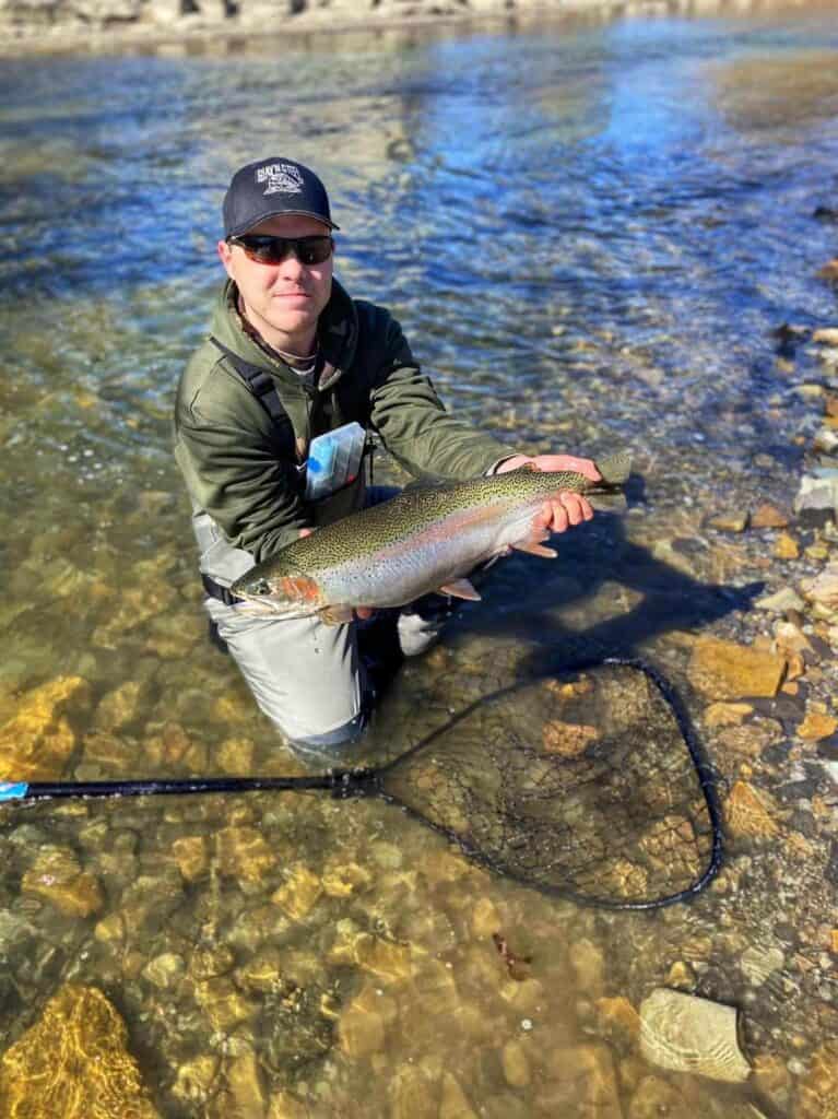 An angler kneels in clear water while holding a large steelhead caught in New York, with a landing net in the stream water in front of him.