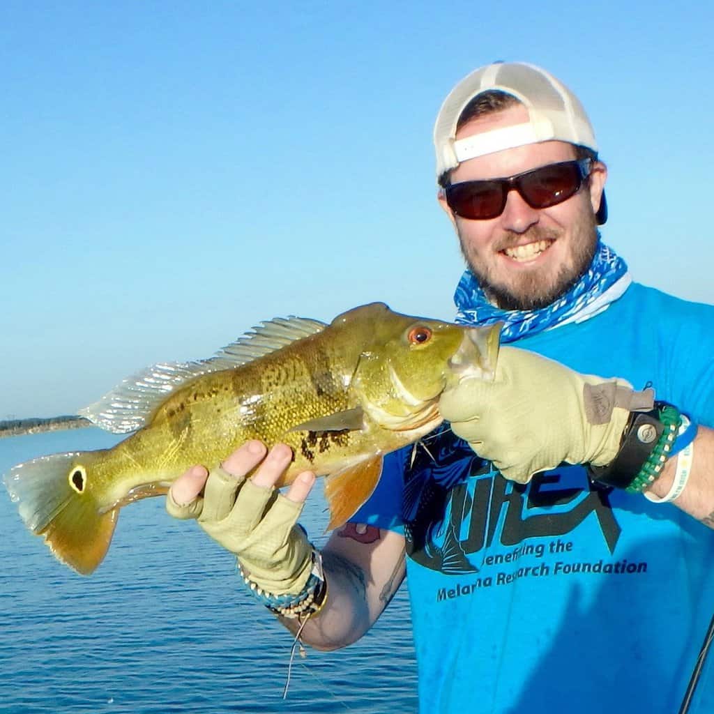 Angler wearing sunglasses and a backwards hat holds a peacock bass he caught fishing in South Florida, with blue sky and water in the background.