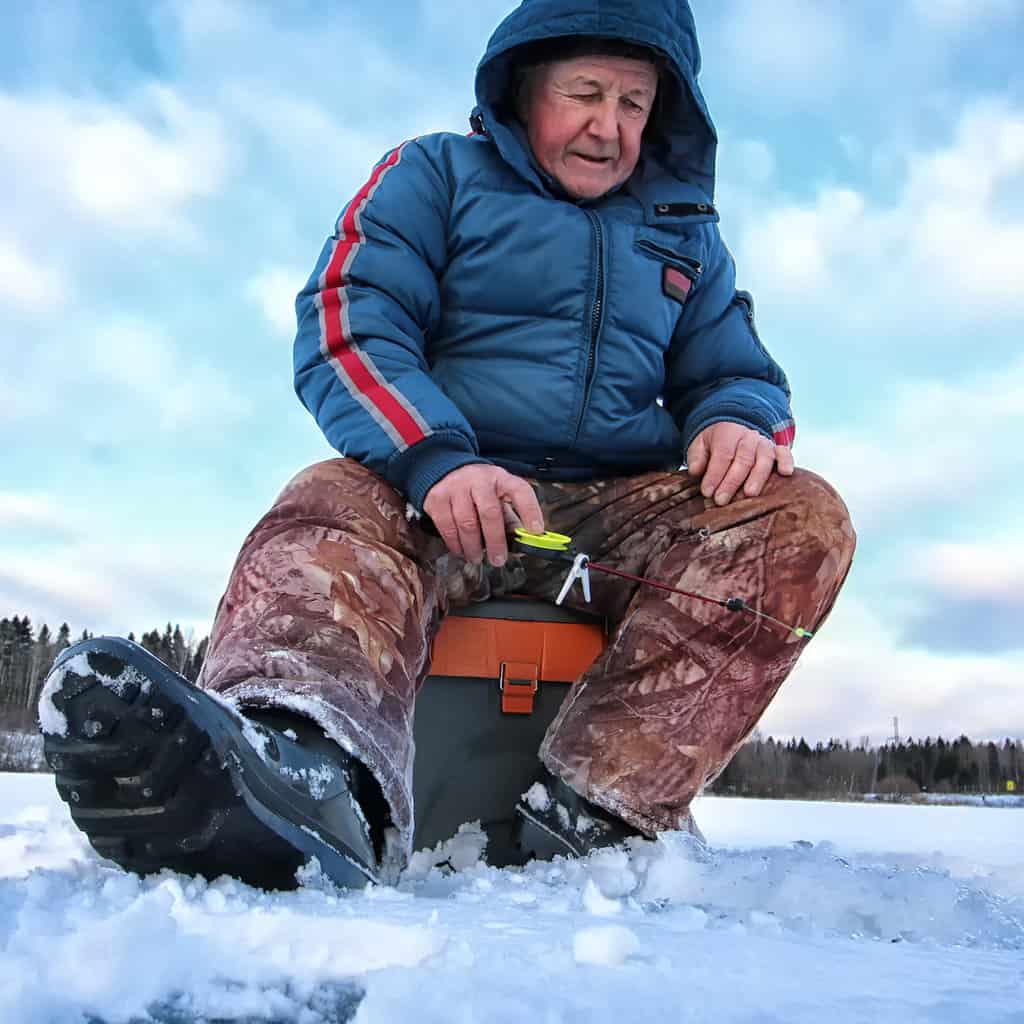 Older man sitting on a tackle box and ice fishing on a frozen lake in New York.