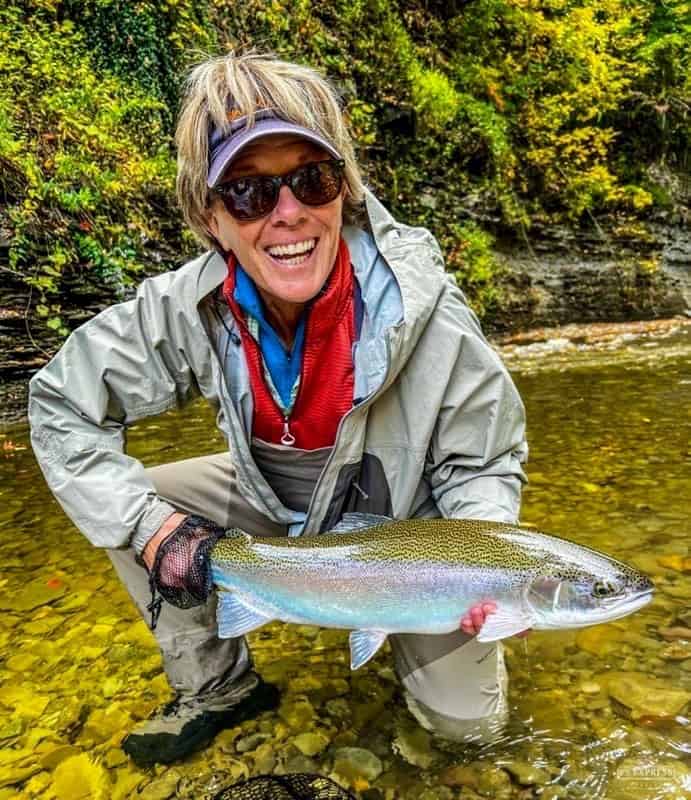 A kneeling and smiling woman holds a beautiful rainbow-colored steelhead with a small creek behind her.