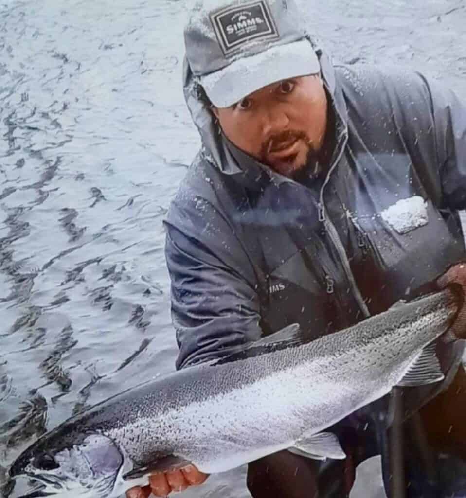 An angler holds a bright silver steelhead caught in cold weather in New York state, with snow stuck to the angler's hat and jacket.