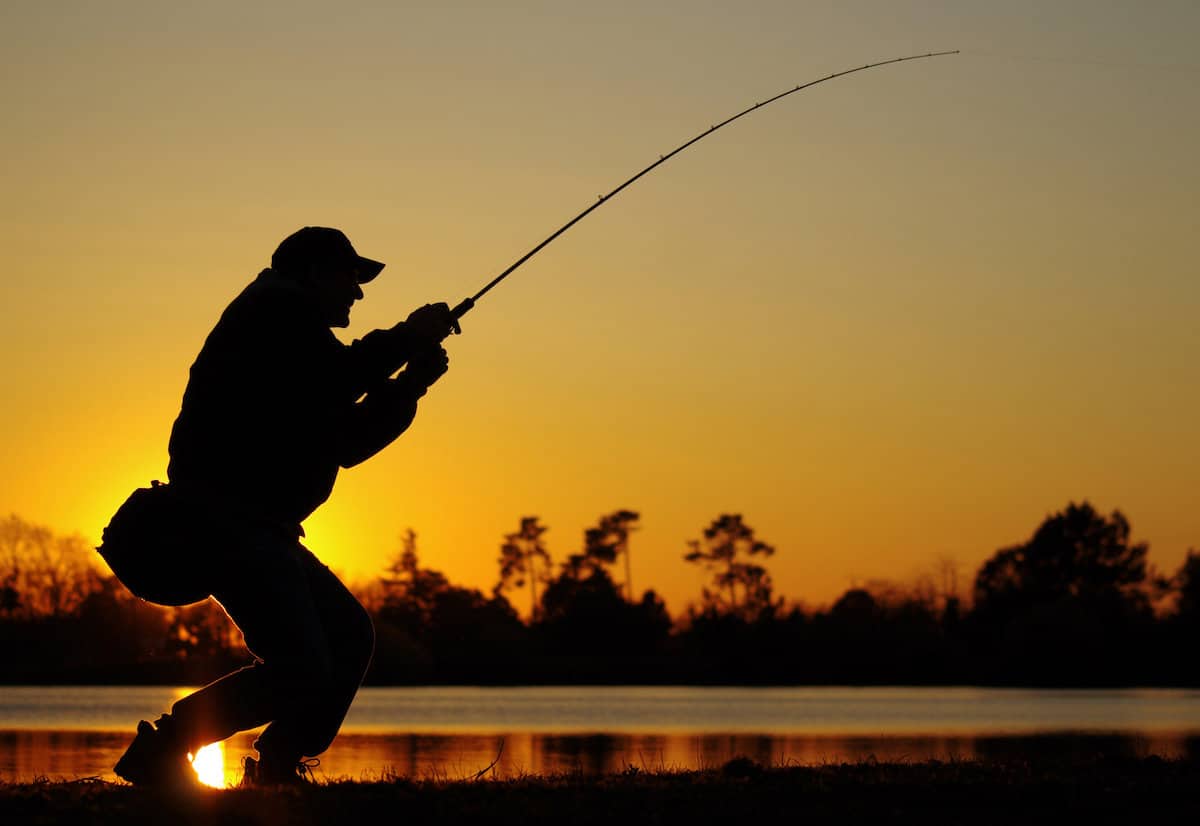An angler fighting a fish in what looks like a good bass lake is shown in silhouette with the setting sun behind his body.
