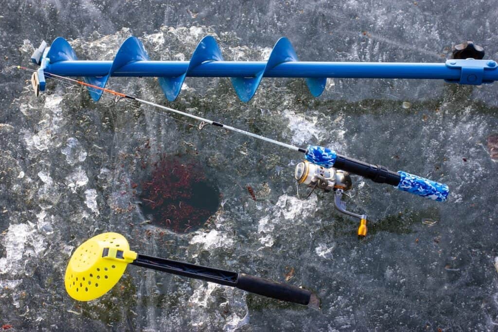 Closeup of ice fishing rod, auger and scoop around a hole in the ice.
