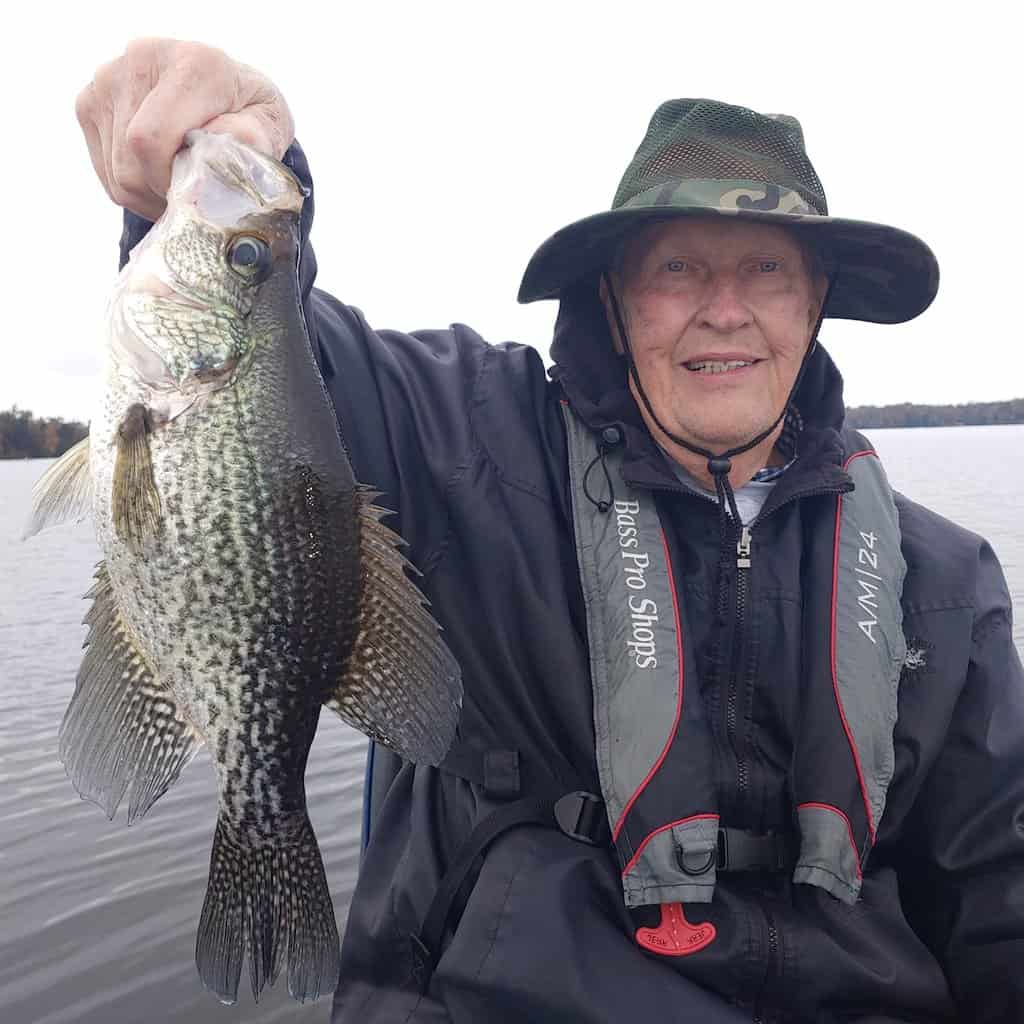 An older angler holds up a large crappie he caught with Lake Talquin, Florida, in the background.