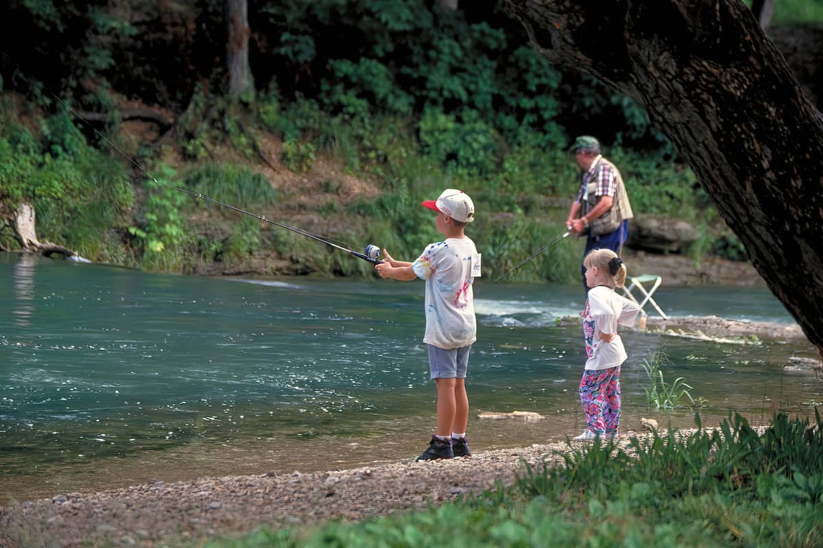 A boy and his grandfather fish for trout in a stream at Roaring River State Park while his sister looks on.