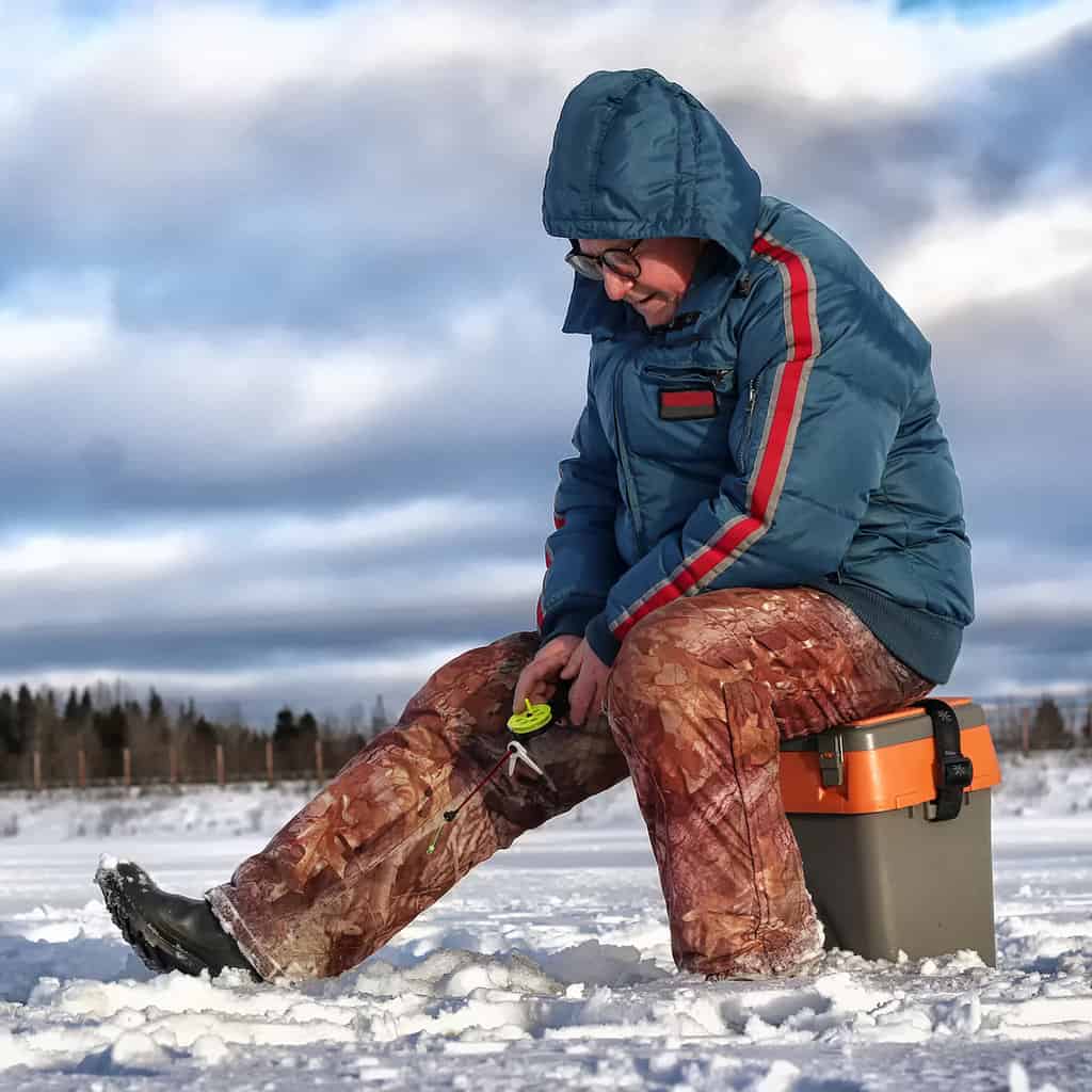 An older man holds an ice fishing pole while sitting on a fishing tackle box on a snow-covered frozen lake in New York state.