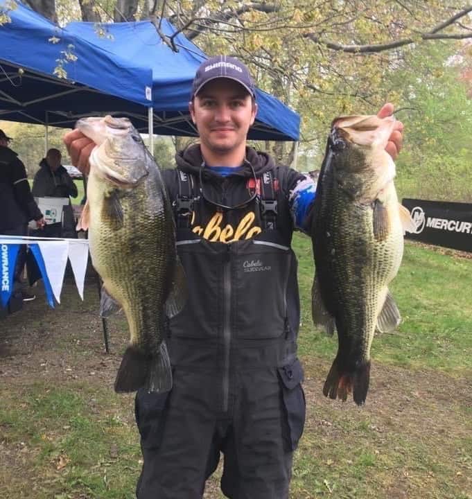 Tournament bass angler Alex Wetherell holds two largemouth bass with bulging bellies, with tournament shelters in the background.