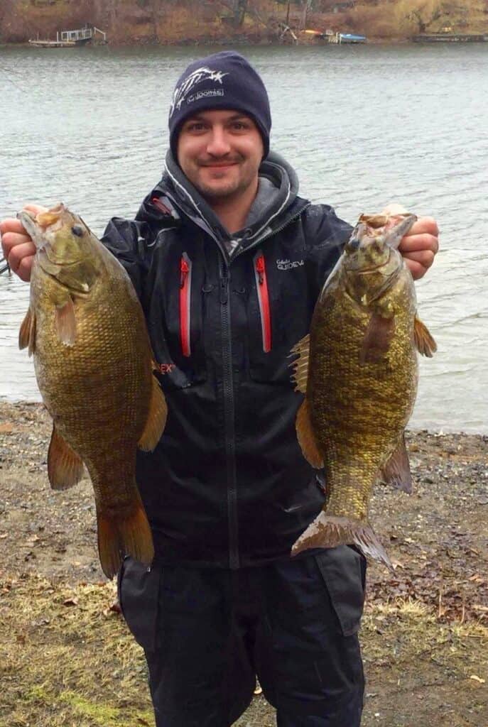 Angler holding two beefy smallmouth bass he caught, with the shoreline of Candlewood Lake right behind him.