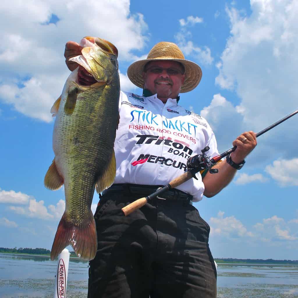 Angler in tournament shirt holds up a big largemouth bass caught in Florida, with a fishing rod in his other hand and a sliver of land and water in the background.