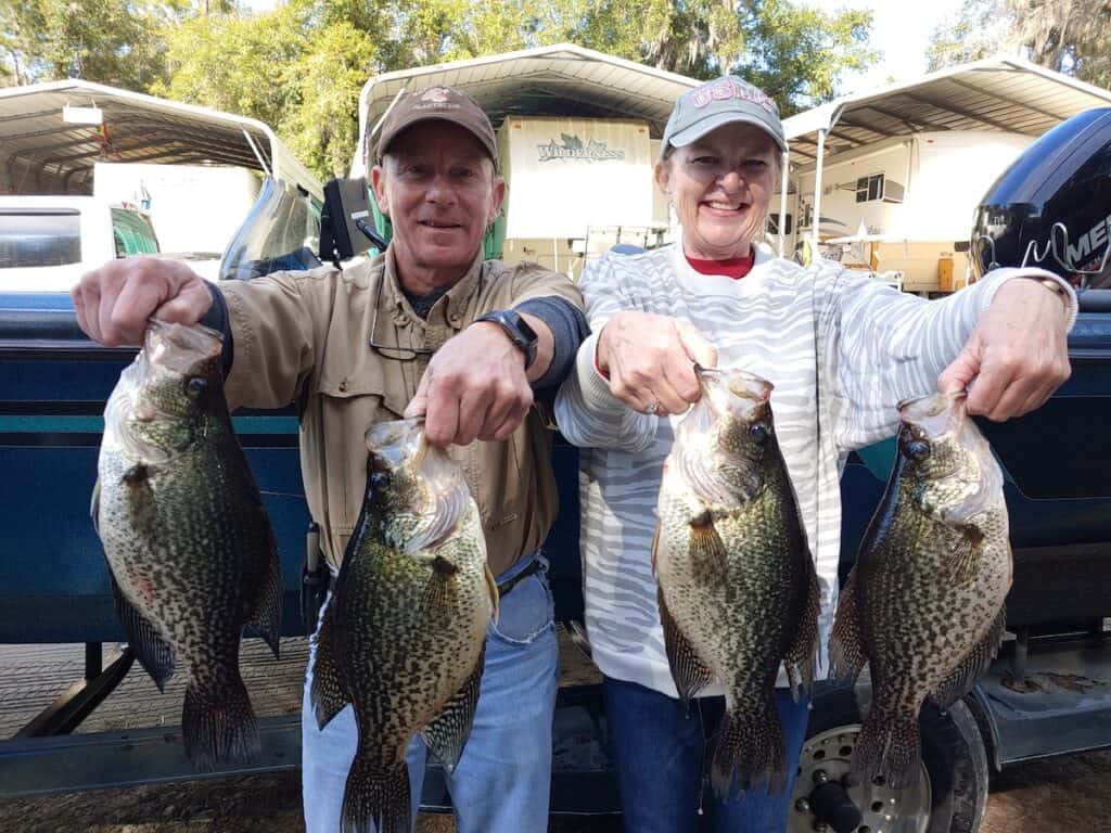A smiling man and woman hold two crappies each after fishing at Lake Talquin, with recreational vehicles behind them.