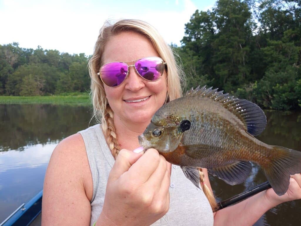 A woman in pink reflective sunglasses holds a large bluegill she caught fishing in Lake Talquin, Florida, which is behind her.