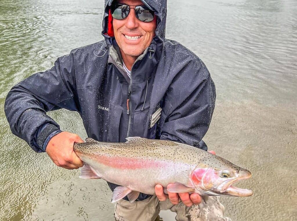 An angler in a blue rain jacket and sunglasses holds a beautifully colored steelhead above the calm waters of Cattaraugus Creek.