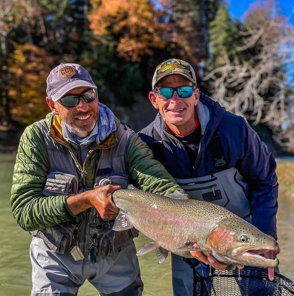 Two male anglers in hats and sunglasses hold a giant steelhead caught fishing in Cattaraugus Creek, New York, which flows in the background.