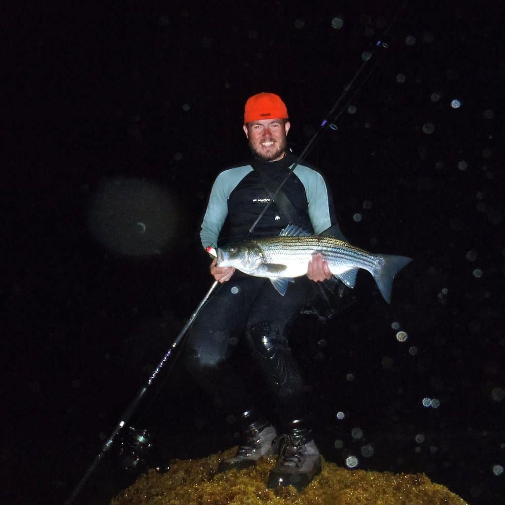 An angler in a red hat holds a striped bass while standing on a rock in Montauk at night.