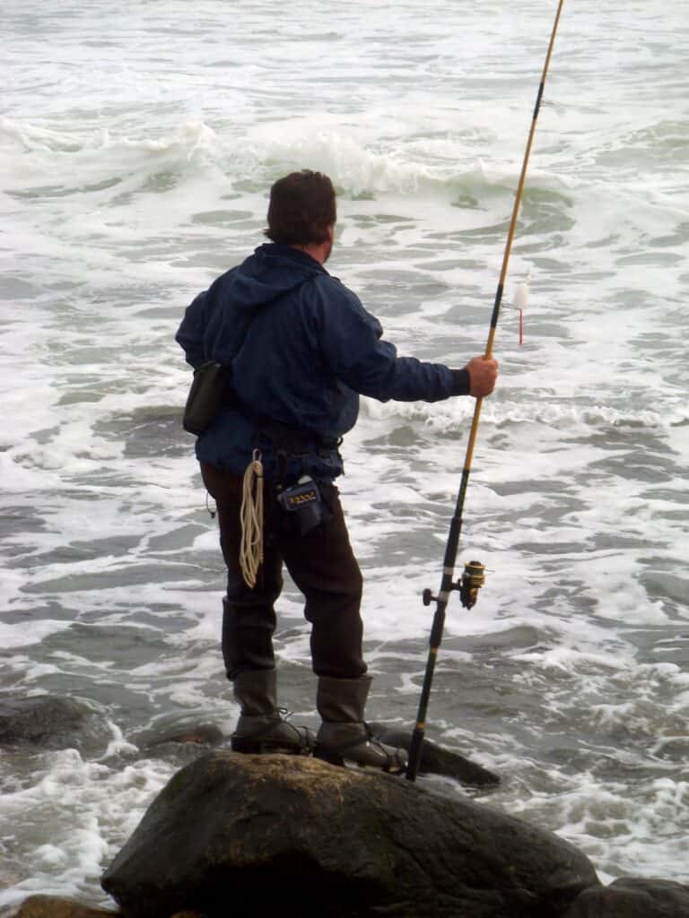 An angler stands on a rock in the surf at Montauk Point while watching the waves and holding a spinning rod, ready to cast for striped bass.