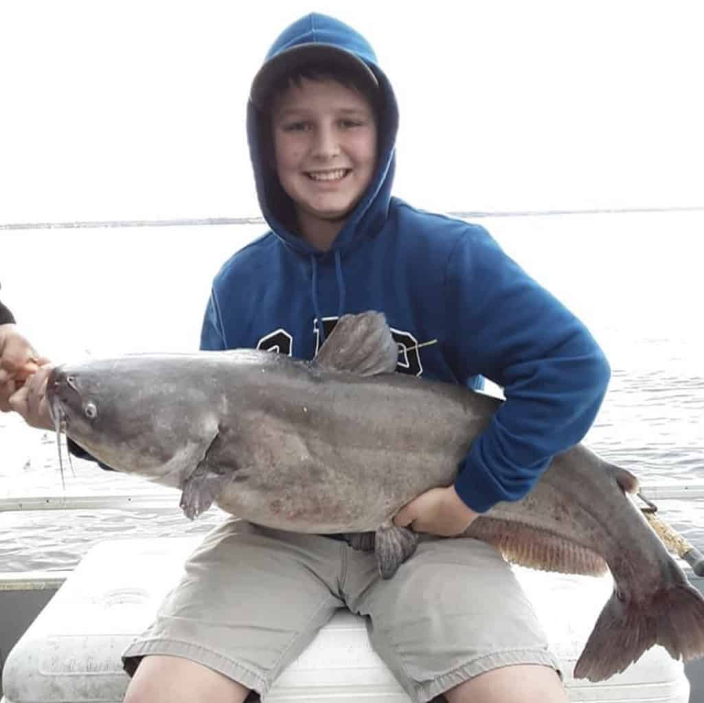 A boy sitting on a boat cooler holds a massive catfish caught fishing in Lake Conroe, Texas, with the lake in the background.