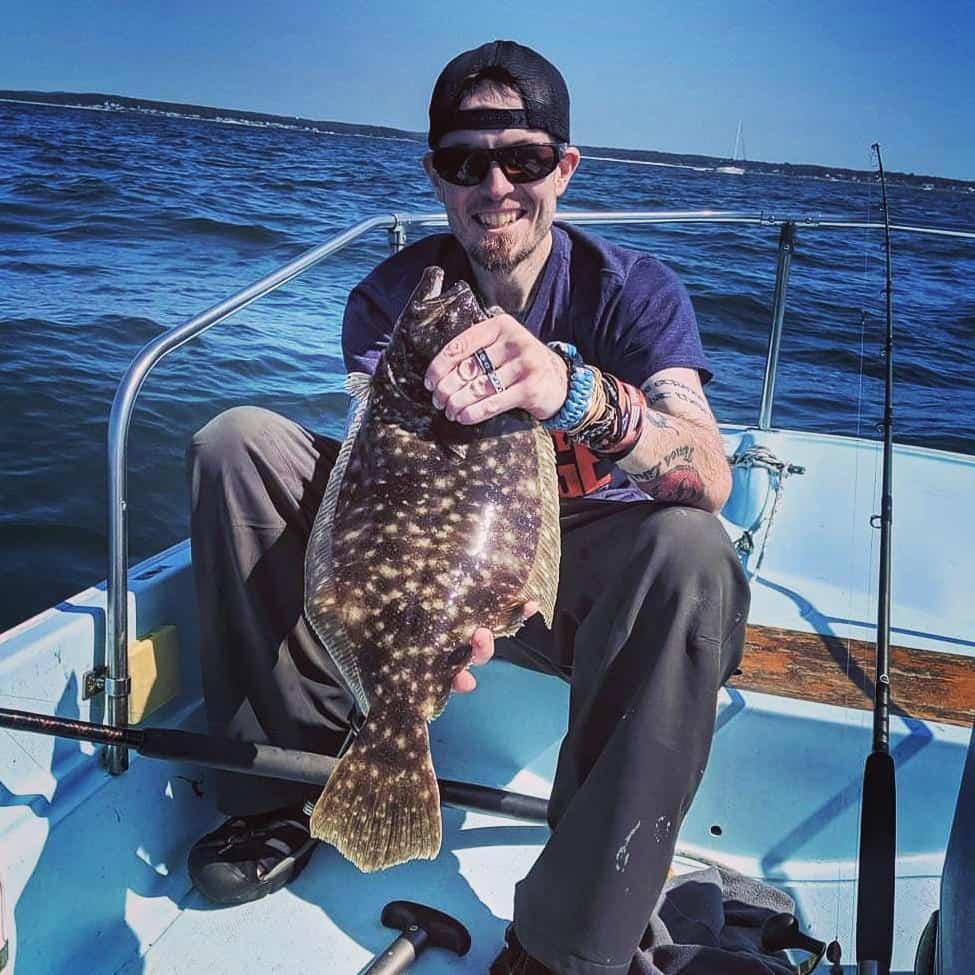 Anglers holds a dark colored flounder while sitting on a fishing boat with the blue Atlantic Ocean in the background.