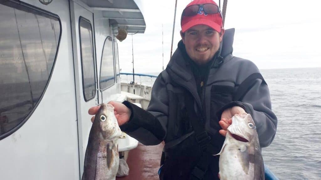 Angler holding two haddock fish while standing on a fishing boat with the Atlantic Ocean behind him.