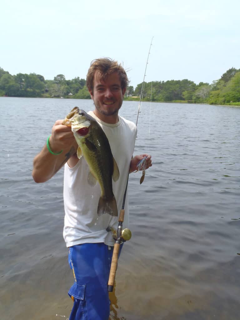 Standing angler holding a largemouth bass caught from one of the many freshwater fishing ponds on Cape Cod, seen behind him.