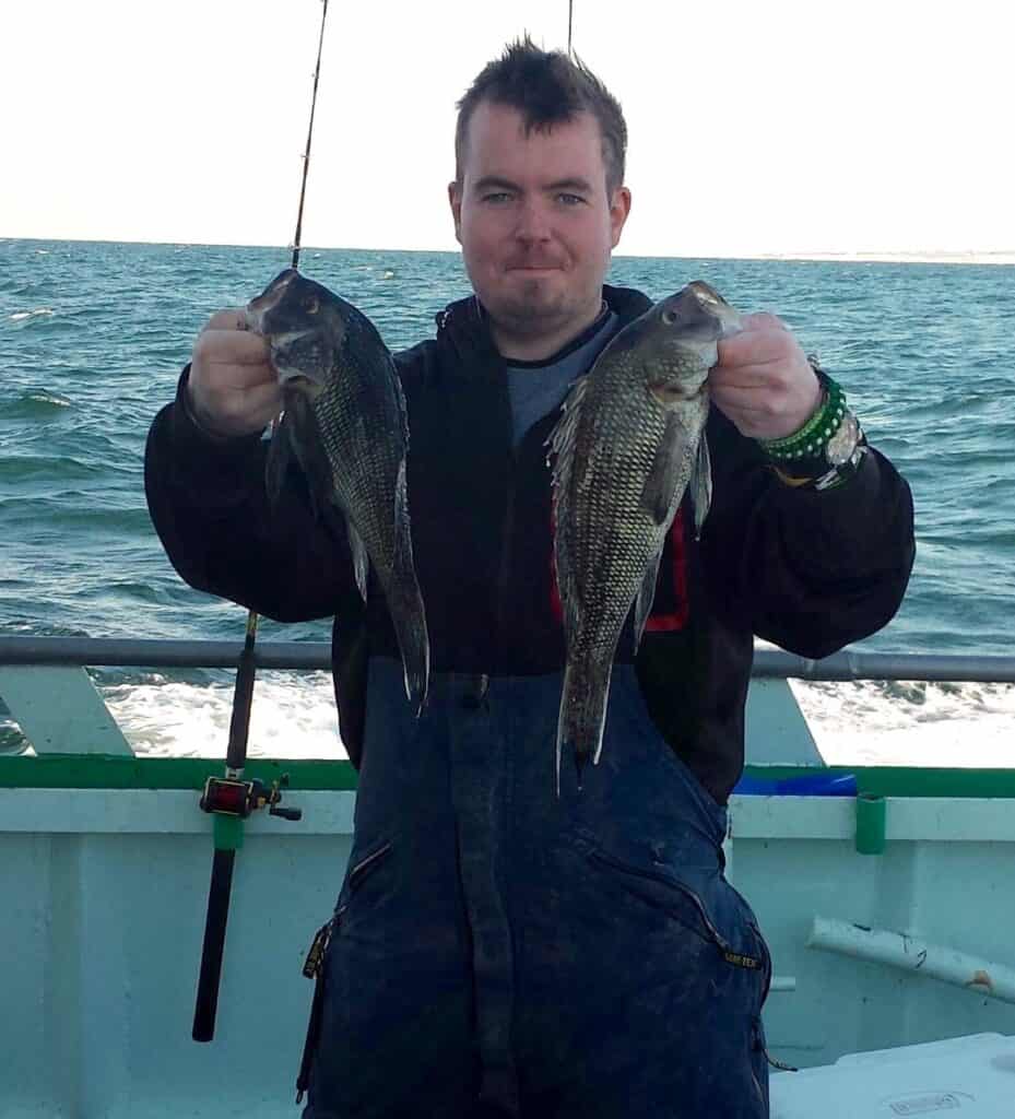 Smiling angler holding two black sea bass with his fishing rod leaning against him and the blue-green water of the Atlantic ocean behind the boat railing.