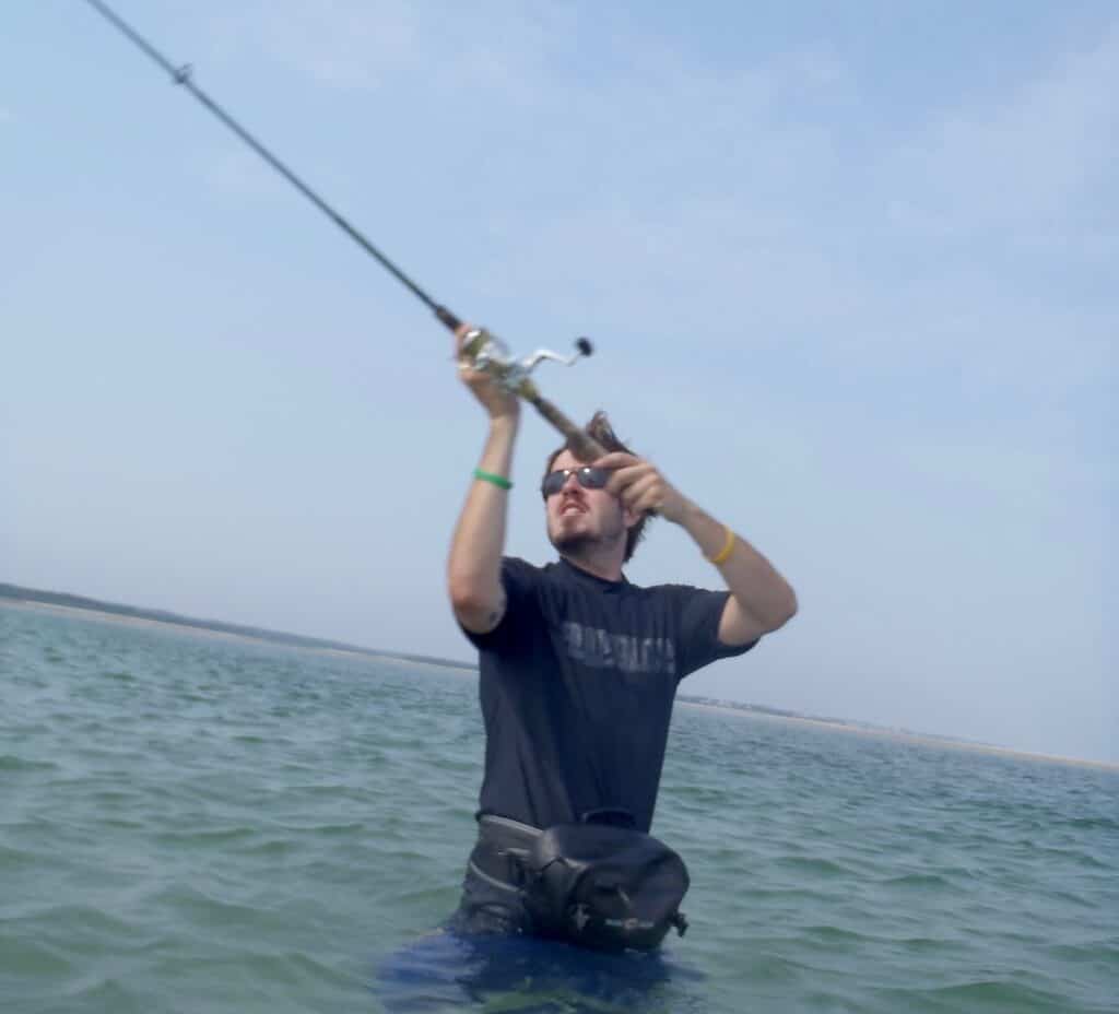 Angler wearing waders and standing in saltwater nearly to his waist preparing to cast while fishing from the shoreline of Cape Cod.