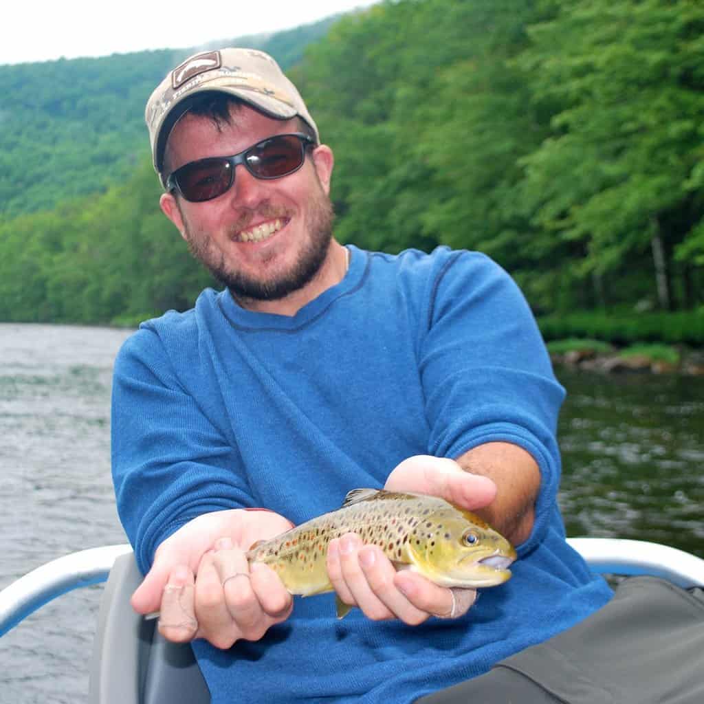 Angler in a boat holds a brown trout caught fishing in the Deerfield River in Massachusetts, with the river and trees in the background.