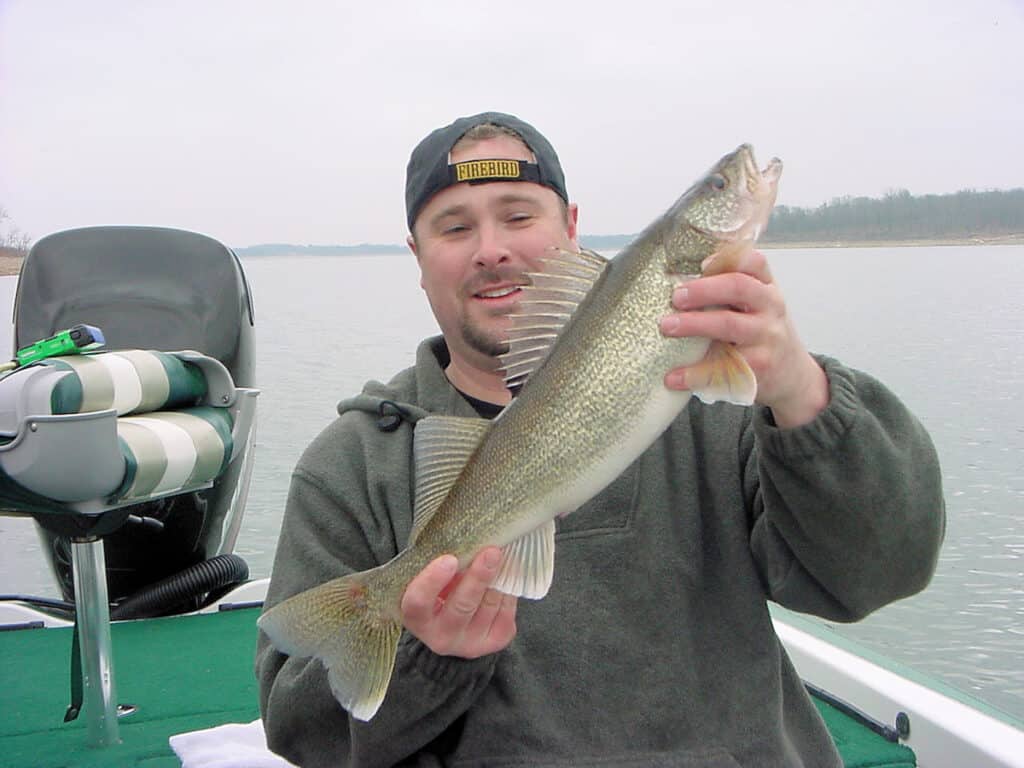 A man wearing a backwards ball cap and sitting in a boat holds up a nice walleye, with Stockton Lake in the background.